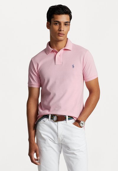 Camisa polo rosa con cuello, mangas cortas y logo en el bolsillo izquierdo del pecho. Combinada con pantalones blancos y un cinturón marrón, con un reloj redondo.