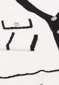 Tessuto lavorato a maglia bianco con strisce nere, scollatura a V e dettagli decorativi con strass sulle alette delle tasche. Superficie con texture.