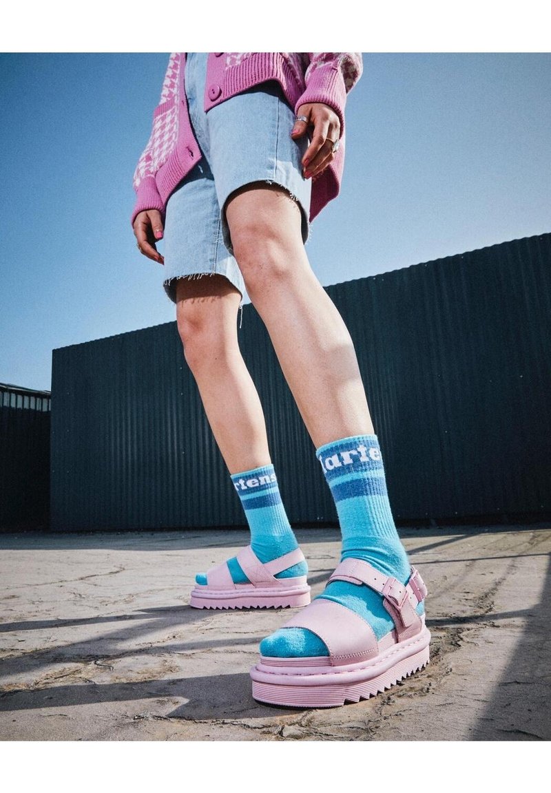 Sandales à plateforme roses avec des brides ajustables, portées avec des chaussettes rayées bleues. Tenue comprenant un cardigan rose et un short en denim. Sol texturé.