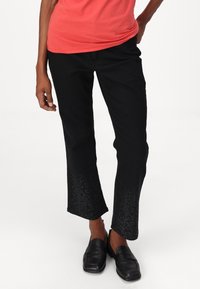 Schwarze Hosen mit strukturierten, glitzernden Details am Saum. Sie haben ein gerades Bein-Design und sind mit schwarzen Slippern kombiniert.