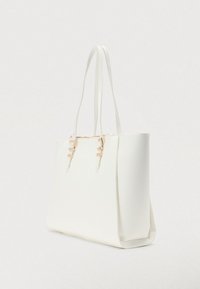 Sac fourre-tout en cuir blanc avec doubles anses d'épaule et accents de boucles dorées, debout sur fond uni.