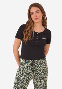 Zwarte top met korte mouwen en een ronde halslijn, met een rij knopen aan de voorkant en een klein geborduurd logo. Gecombineerd met bloemenbroek.