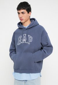 Temno modra eveni pulover z motivom »GAP Athletic« v beli barvi, žepom vrsta kenguru in ohlapnim krojem, nošen prek svetlo modre črtaste srajce.