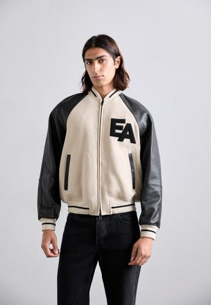 Ung mand med skulderlangt hår iført en beige og sort varsityjakke med "EA" på brystet og sorte jeans mod en grå baggrund.