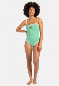 Ολόσωμο μαγιό σε μέντα πράσινο με λεπτές λευκές τιράντες, διαθέτει μικρό πολύχρωμο λογότυπο στο μπροστινό μέρος, εφαρμοστή γραμμή, λεία υφή.