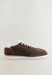 Zapatilla deportiva marrón que presenta una combinación de materiales de ante y nylon, acentos blancos, suelas texturizadas y un diseño clásico de cordones.