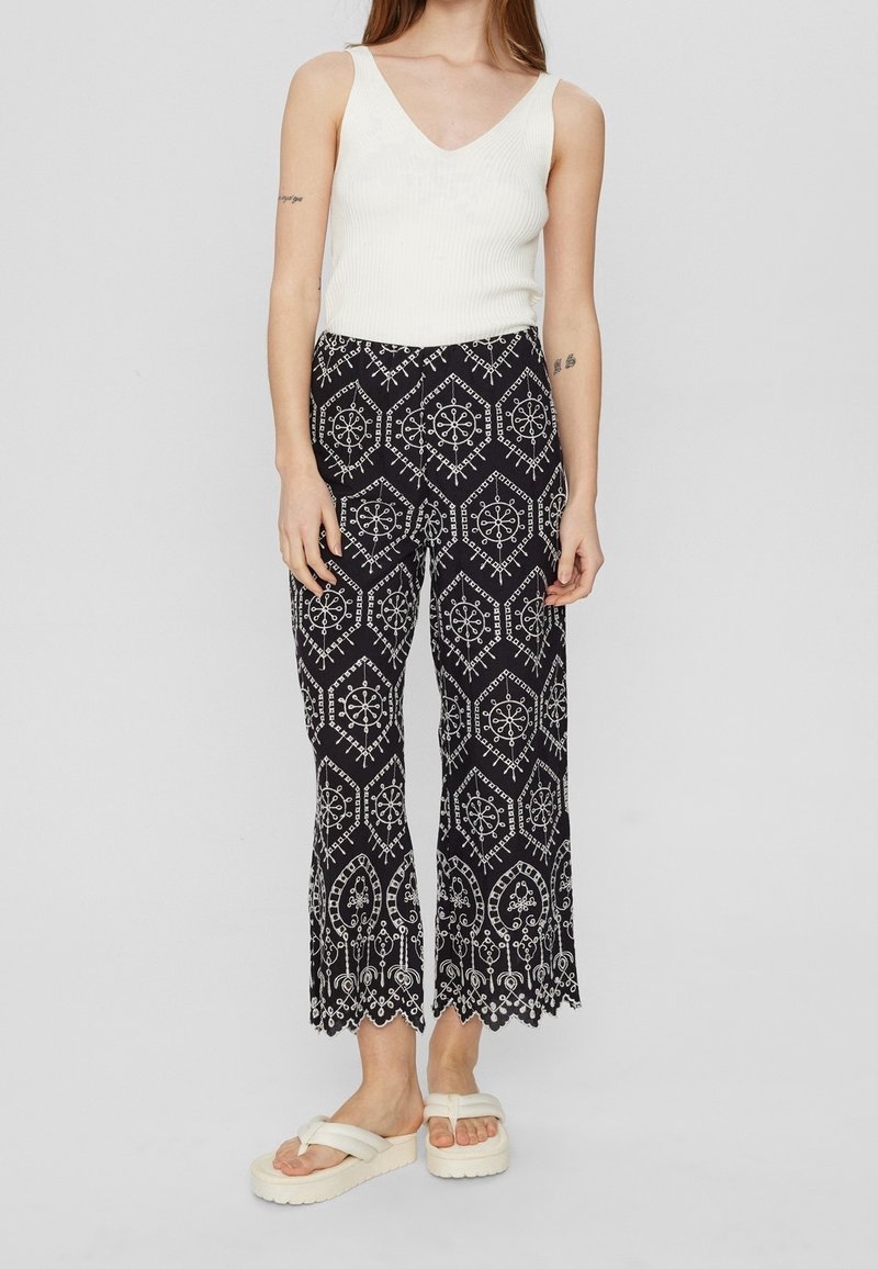 Jeune femme portant un débardeur blanc, un pantalon large noir avec des motifs géométriques blancs et des sandales compensées blanches.