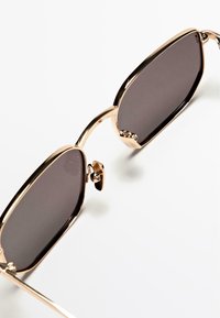 Zonnebril met een metalen montuur in goudkleur, donkere grijze glazen en een slank design. Voorzien van neuspads voor comfort en een afgeronde vorm.