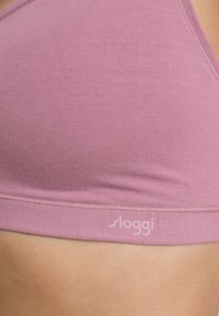 Bralette rose clair en tissu doux avec une texture lisse. Doté d'une bande étroite et d'un petit logo "sloggi" sur le côté gauche.