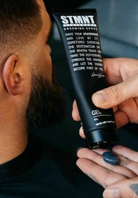 Mann gibt dunkeltgraues Pflegemittel aus einer schwarzen STMNT-Tube in die Handfläche ab, Nahaufnahme der Hand, des Produkts und der Seite des Bart tragenden Mannes.