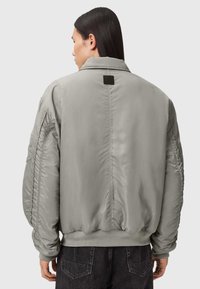 Chaqueta bomber gris con una textura suave y lisa, puños y cinturilla elásticos. Presenta una costura y una etiqueta rectangular en la parte posterior.