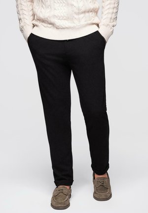 Homme portant un pantalon noir avec les mains dans les poches, un pull en tricot de crème et des chaussures en daim marron à lacets, se tenant devant un fond uni.