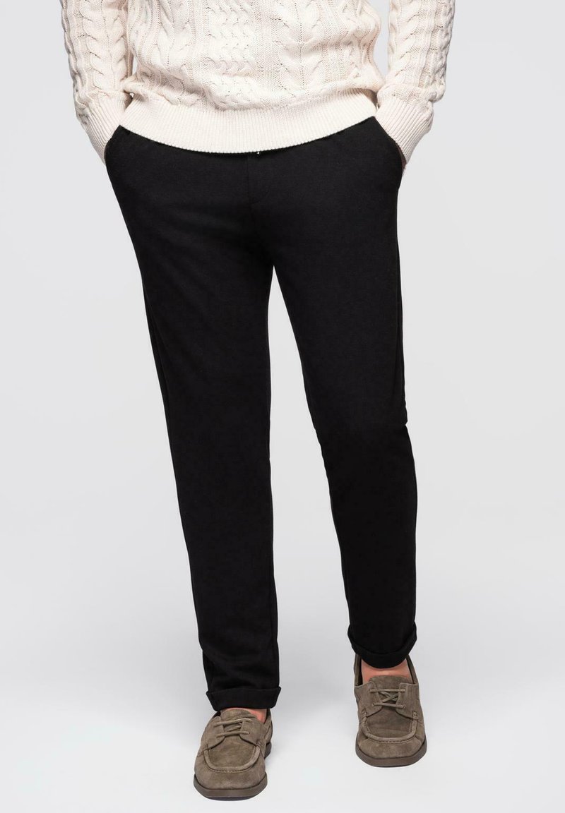 Homme portant un pantalon noir avec les mains dans les poches, un pull en tricot de crème et des chaussures en daim marron à lacets, se tenant devant un fond uni.