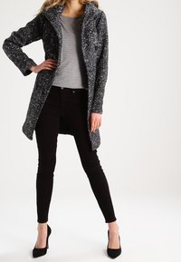 Grå och svart prickig kappa med huva, knapplist fram, och två fickor, i kombination med en grå topp och svarta smala byxor.