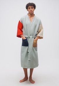 Άνδρας που στέκεται ξυπόλητος, φορώντας μια υφασμάτινη ανοιχτό γκρι ρόμπα με κόκκινα και μπεζ μανίκια και μπροστινές τσέπες σε σκούρο μπλε και καφέ χρώμα.