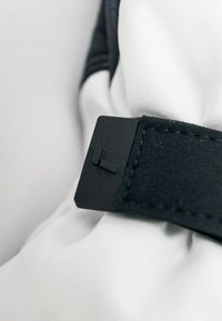 Étiquette rectangulaire noire attachée à une sangle en tissu blanc, avec une surface texturée et une lettre en relief subtile. Design simple avec des couleurs neutres.