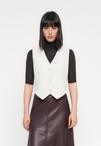 Chaleco blanco ajustado con escote en forma de V y tres botones. Llevado sobre un cuello de tortuga de rayas oscuro, combinado con una falda de cuero burdeos.