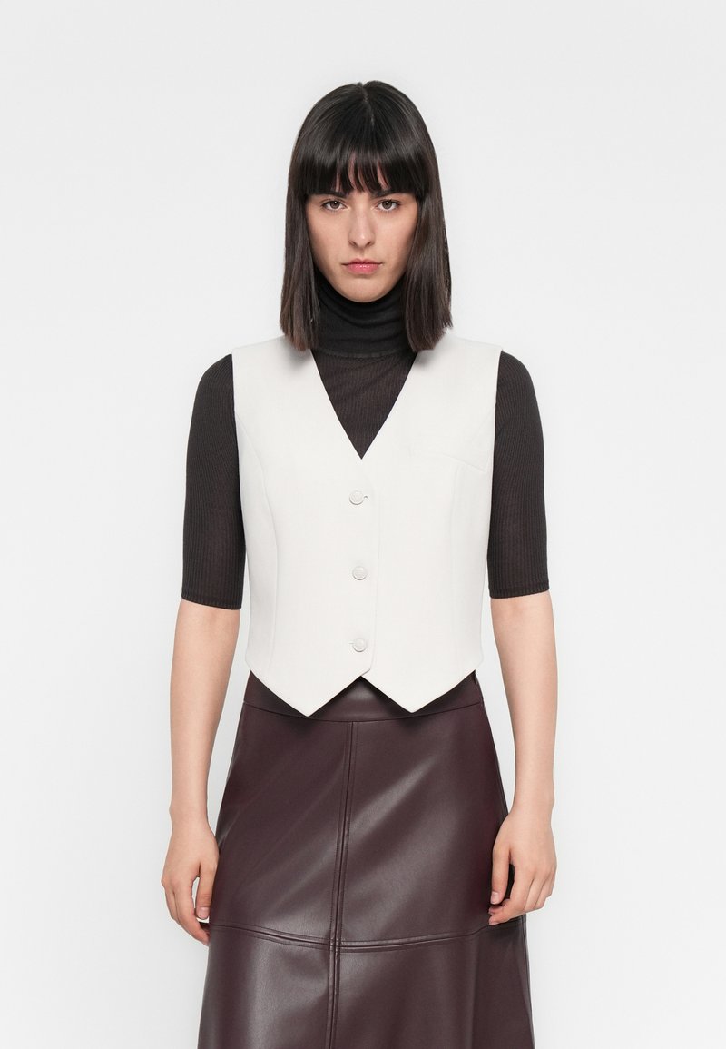 Chaleco blanco ajustado con escote en forma de V y tres botones. Llevado sobre un cuello de tortuga de rayas oscuro, combinado con una falda de cuero burdeos.