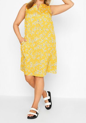 Keltainen hihaton mekko, jossa on kukkakuvio ja V-aukko sekä sivutaskut. Yhdistetty valkoisiin ja mustiin sandaaleihin.