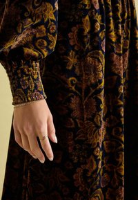 Dunkelblumiges Samtkleid mit reichhaltigen Mustern in Gold, Burgunder und Marineblau. Geraffte Ärmelbündchen und ein fließendes Design betonen den Stoff.