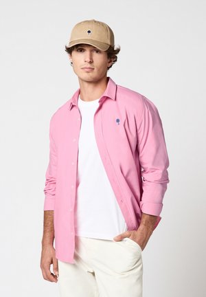 Jeune homme portant une casquette beige, une chemise rose boutonnée sur un t-shirt blanc, et un pantalon clair, debout avec une main dans la poche sur un fond uni.