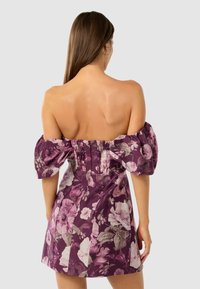 Vrouw met lang bruin haar in een paarse off-shoulder jurk met pofmouwen en bloemenmotief, van zich af gekeerd tegen een effen achtergrond.