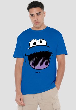 Helles blauer T-Shirt mit einem breiten Munddesign eines Cartoon-Charakters in Schwarz und Lila, mit großen weißen Augen über dem Mund.