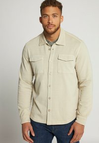 Camisa beige de manga larga con dos bolsillos en el pecho, cierre de botones en el frente y tejido ligeramente texturizado, llevada sobre una camisa gris con estampado.
