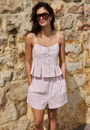 Femme aux longs cheveux foncés et grandes lunettes de soleil, portant un haut sans manches à rayures roses avec des boutons et un short assorti, debout contre un mur en pierre.