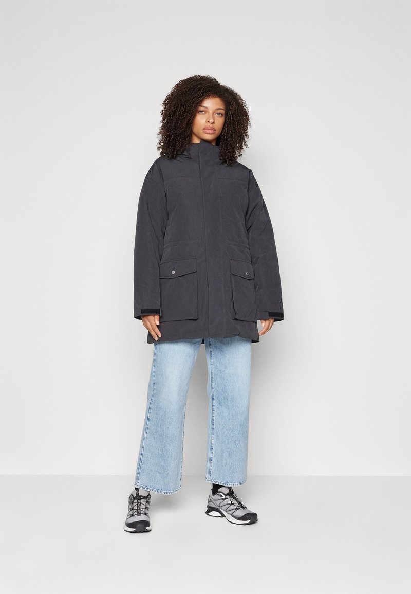 Parka noire avec un col montant, deux poches avant et une coupe ample, associée à un jean large bleu clair et des baskets grises.