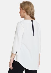 Weiße Bluse mit Dreiviertelärmeln, einem abgerundeten Saum und einem marineblauen Reißverschlussdetail auf der Rückseite. Glattes Material mit subtiler Struktur.