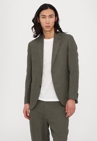 Olive green blazer with a textured weave, single-breasted design, and two buttons. Paired with a white t-shirt and matching trousers.