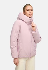 Vrouw in een lichtroze gewatteerde jas met hoge kraag, die de rits vlakbij haar gezicht vasthoudt, gekleed in crèmekleurige broek, tegen een effen achtergrond.