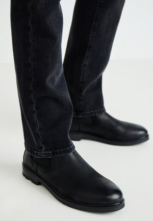 Botas Chelsea de cuero negro usadas con vaqueros ajustados gris oscuro, de pie sobre una superficie blanca.