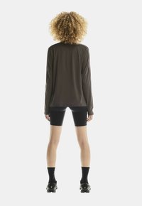 Persoon met krullend haar staat met de rug naar de camera, gekleed in een zwart shirt met lange mouwen, zwarte fietsshorts, zwarte sokken en zwarte schoenen.
