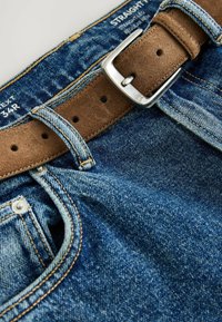 Ceinture en daim marron avec boucle en argent enfilée dans les passants d'un jean en denim bleu étiqueté taille 34R avec une étiquette coupe droite.