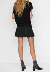 Schwarzes Kurzarmoberteil kombiniert mit einem gefalteten schwarzen Rock. Das Modell trägt zudem graue Socken und glänzende schwarze High Heels mit Blockabsatz.