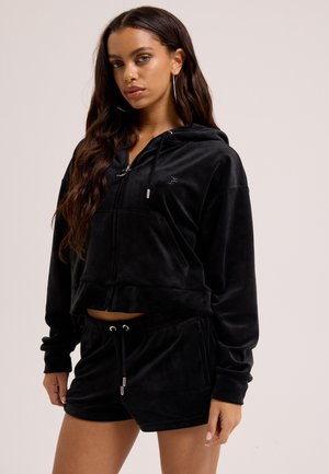 Ung kvinde med langt hår iført sort velour-hættetrøje og matchende shorts mod en ensartet lys baggrund.