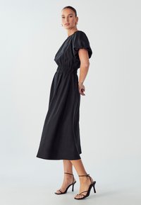 Črna midi obleka s pufastimi rokavi in zategnjenim pasom. Gladka tkanina, ravna silhueta. V kombinaciji s črnimi visokimi petami z naramnicami.