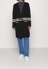 Schwarzer Mantel mit Kapuze und weißen Streifen an der Taille und den Ärmeln, ausgestattet mit zwei Fronttaschen und einem gebundenen Gürtel. Kombiniert mit blauen Jeans und weißen Schuhen.