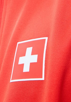Czerwony materiał z białym symbolem krzyża w czerwonym kwadracie, przypominający emblem pierwszej pomocy lub szwajcarską flagę na ubraniu.