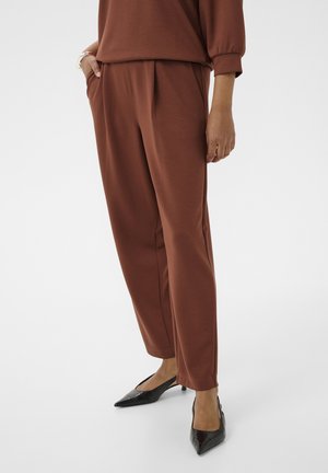 Persona vistiendo pantalones marrones de corte suelto y top a juego con zapatos kitten de punta negra, de pie con una mano en el bolsillo.