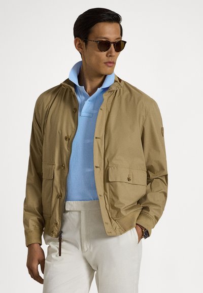 Ανοιχτό καφέ μπουφάν με φερμουάρ μπροστά, δύο τσέπες στο στήθος και ριμπ μανσέτες, φορεμένο πάνω από ανοιχτό μπλε πόλο, σε συνδυασμό με λευκό παντελόνι.