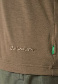 Tissu marron avec logo VAUDE brodé et petite étiquette verte portant la mention « GREEN SHAPE » sur la couture latérale au-dessus d'un pantalon vert.