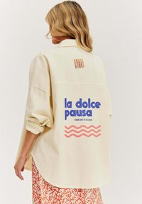 Krémová oversized košeľa s textom "la dolce pausa" v modrej farbe, grafickými vlnami v ružovej a kockovanou nášivkou na zadnej strane. Bavlnený materiál.