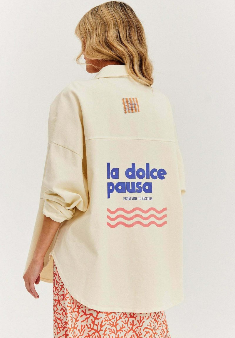 Krémová oversized košeľa s textom "la dolce pausa" v modrej farbe, grafickými vlnami v ružovej a kockovanou nášivkou na zadnej strane. Bavlnený materiál.