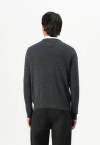 Uomo in maglione grigio scuro a maniche lunghe e pantaloni neri, in piedi di spalle contro uno sfondo bianco e semplice.