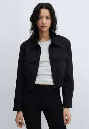 Black cropped jacket in sturdy fabric, featuring a pointed collar, two chest pockets, and elasticated hem. Worn over a grey top.
