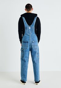 Denim overalls in light blue, featuring a loose fit, adjustable shoulder straps, large front pockets, and contrast stitching. Black hoodie underneath.