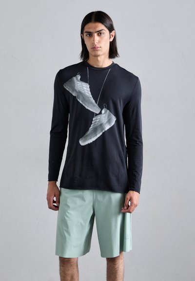 Schwarzes langärmliges rippiertes T-Shirt mit einem grafischen Druck von Schuhen. Kombiniert mit hellgrünen Shorts, die ein kontrastierendes Outfit schaffen.